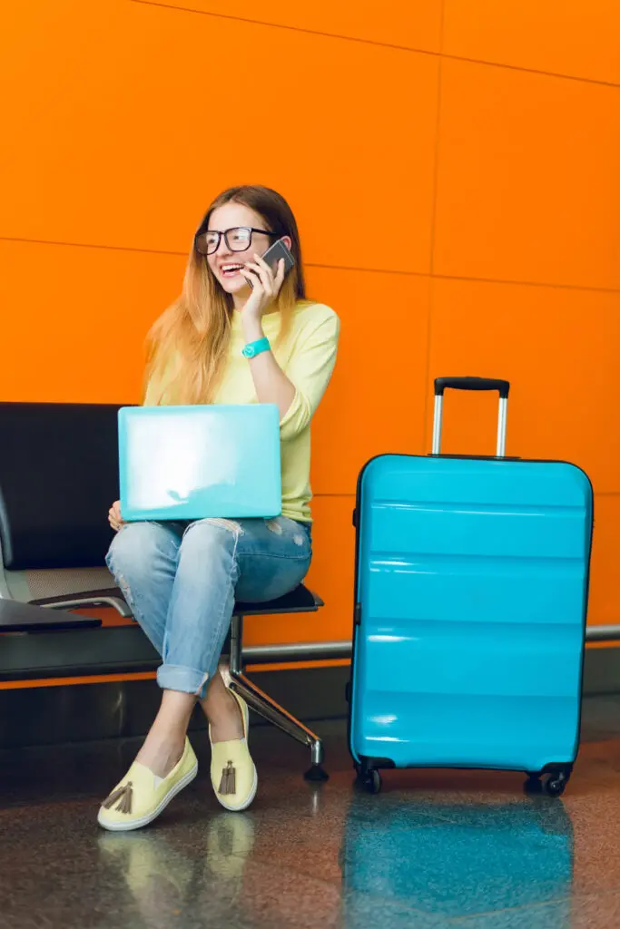 pretty girl with long hair is sitting on chair on orange background. she wears jeans with jellow sweater. she has laptop and suitcase near. she is speaking on phone